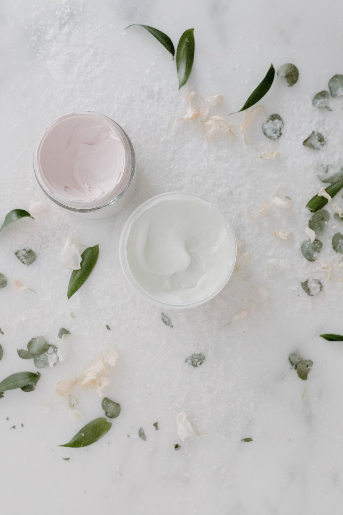 Two open cream jars on marble surrounded by leaves and petals